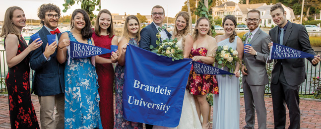 Bride and groom stand behind Brandeis banners surrounded by people, several in floral dresses